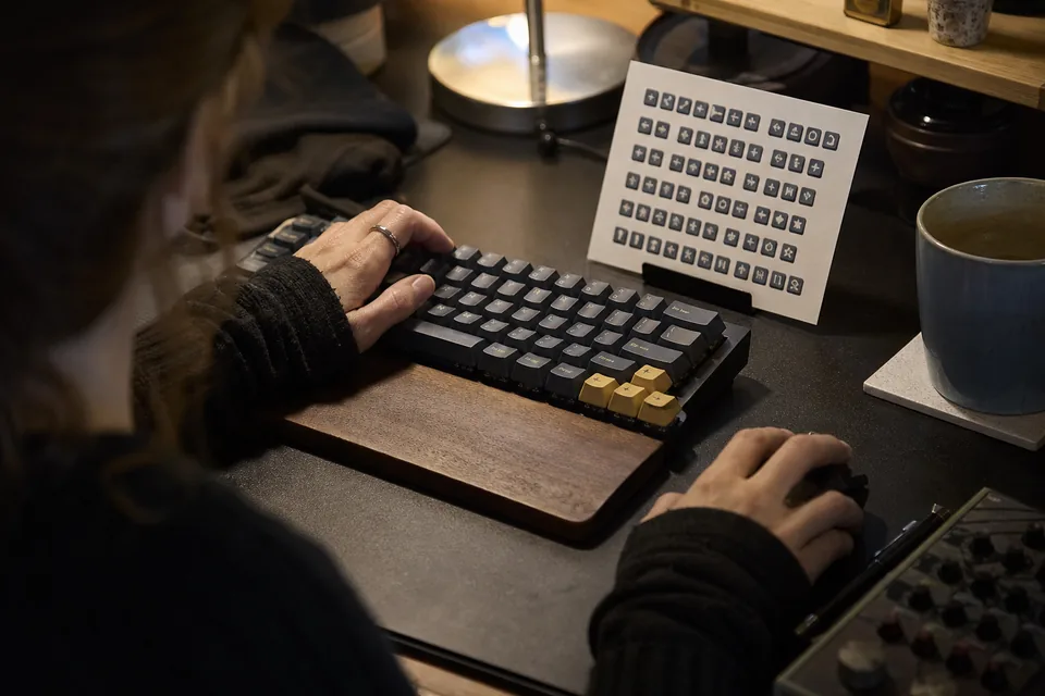 Close view of hands on a compact keyboard and a mouse at a labeling desk with a printed shortcut card